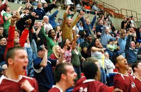 A large, diverse group of cheering students, standing up and fist-pumping on the bleachers of a basketball game